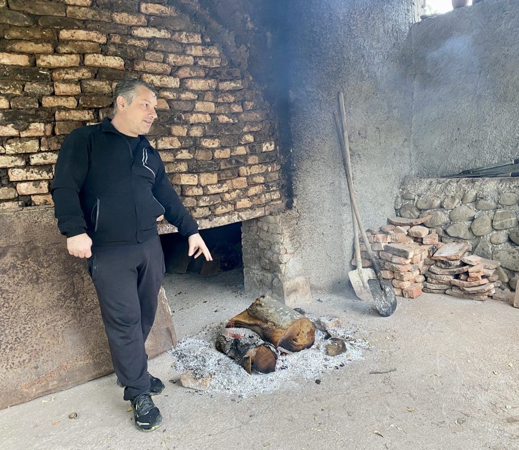 A qvevri maker pointing to wood in front of a stone structure